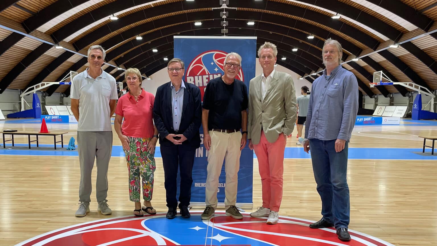Sechs Personen posieren in der Sporthalle der RheinStars Köln vor dem Teamlogo.