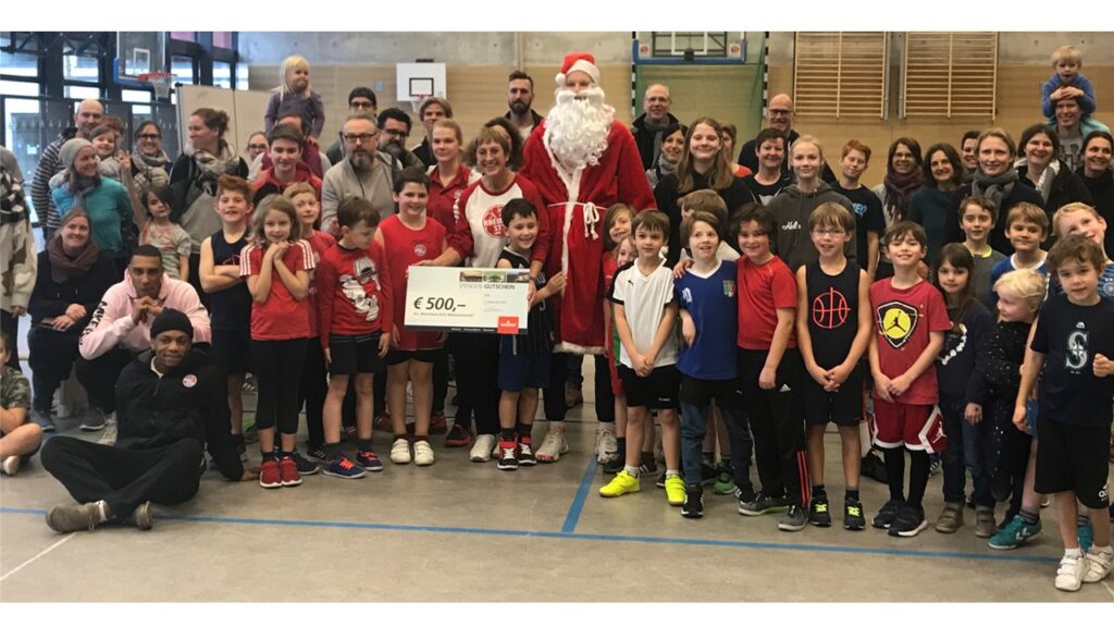 Große Gruppe von Kindern und Erwachsenen mit dem Weihnachtsmann in einer Sporthalle, hält einen Scheck über 500 Euro.