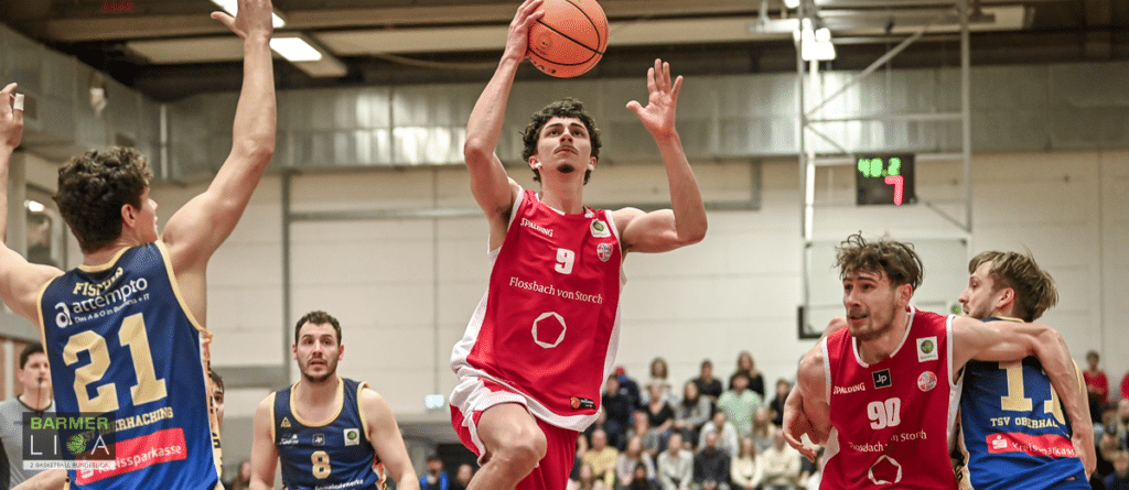 Basketballspieler in rotem Trikot wirft Ball während Spiel gegen Team in blauem Trikot.