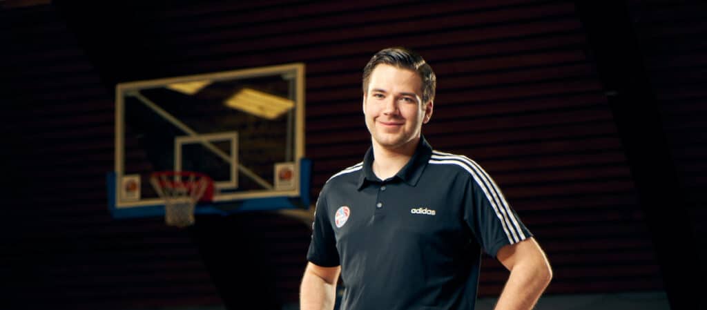 Ein Mann in schwarzem Adidas-Poloshirt steht lächelnd vor einem Basketballkorb in einer RheinStars Köln-Halle.