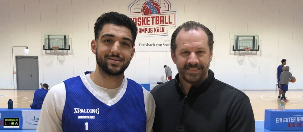 Zwei Männer posieren in einer Basketballhalle der RheinStars Köln, einer trägt das blaue Trikot Nummer 1.