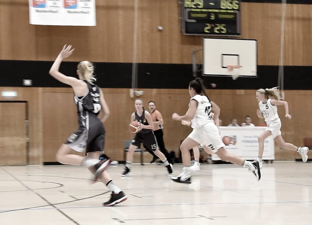 Basketballspiel der RheinStars Köln in der Halle: Spielerin in schwarzem Trikot springt zum Wurf, während Gegnerinnen in weißen Trikots verteidigen.