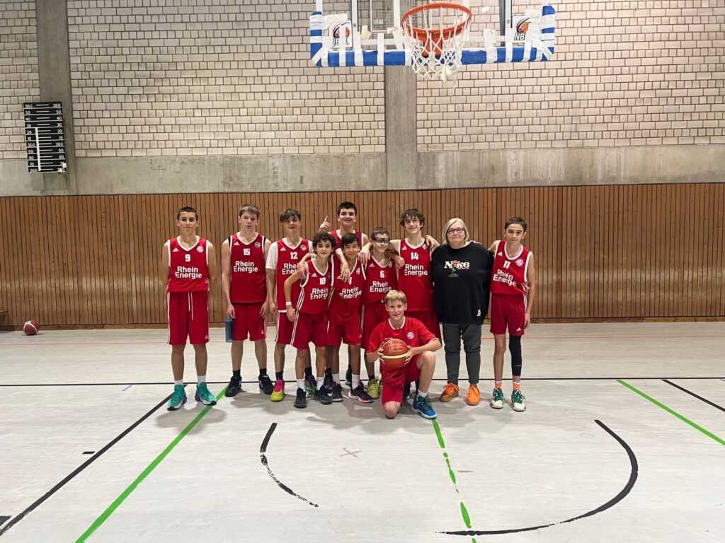 Basketballmannschaft RheinStars Köln posiert in roter Trainingskleidung in einer Sporthalle.