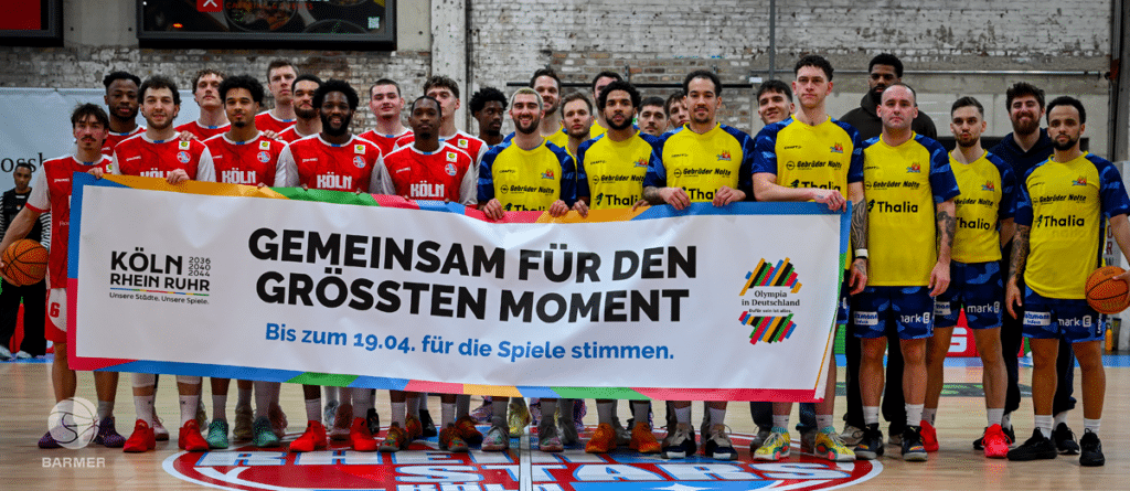 Zwei Basketballmannschaften halten ein Wahlplakat der RheinStars Köln in einer Sporthalle.