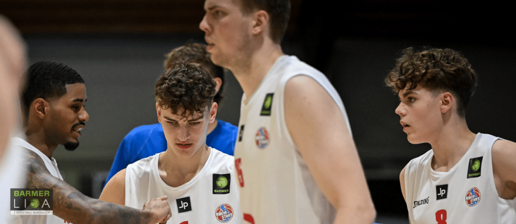 Basketballspieler der RheinStars Köln in weißen Trikots während einer Spielpause im Gespräch.