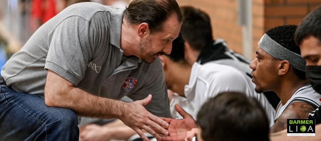 Ein Mann in grauem Polo-Shirt spricht mit einem Basketballspieler der RheinStars Köln während eines Trainings.