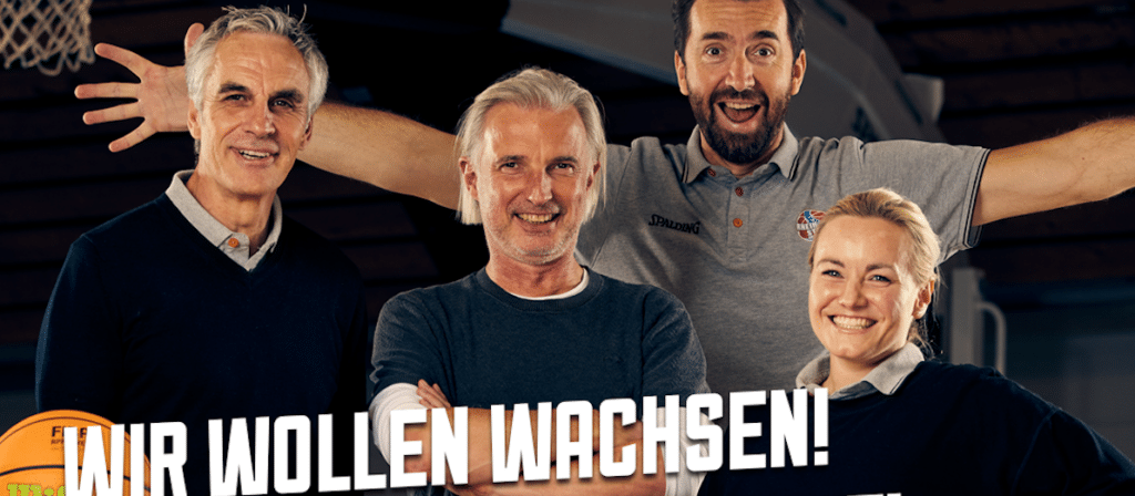 Vier lächelnde Personen posieren zusammen vor einer Basketballanlage mit dem Text "Wir wollen wachsen!" und dem RheinStars Köln Logo.