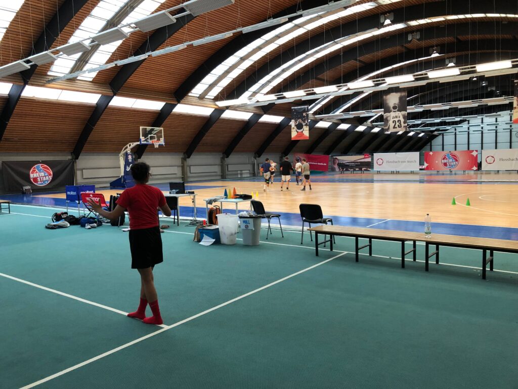 Trainingshalle der RheinStars Köln mit Basketballplatz, Spielern und Trainingsausrüstung.