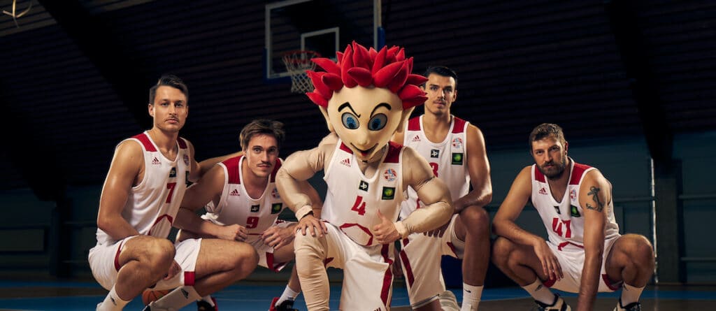 Fünf männliche Basketballspieler der RheinStars Köln posieren mit ihrem Maskottchen in der Sporthalle.