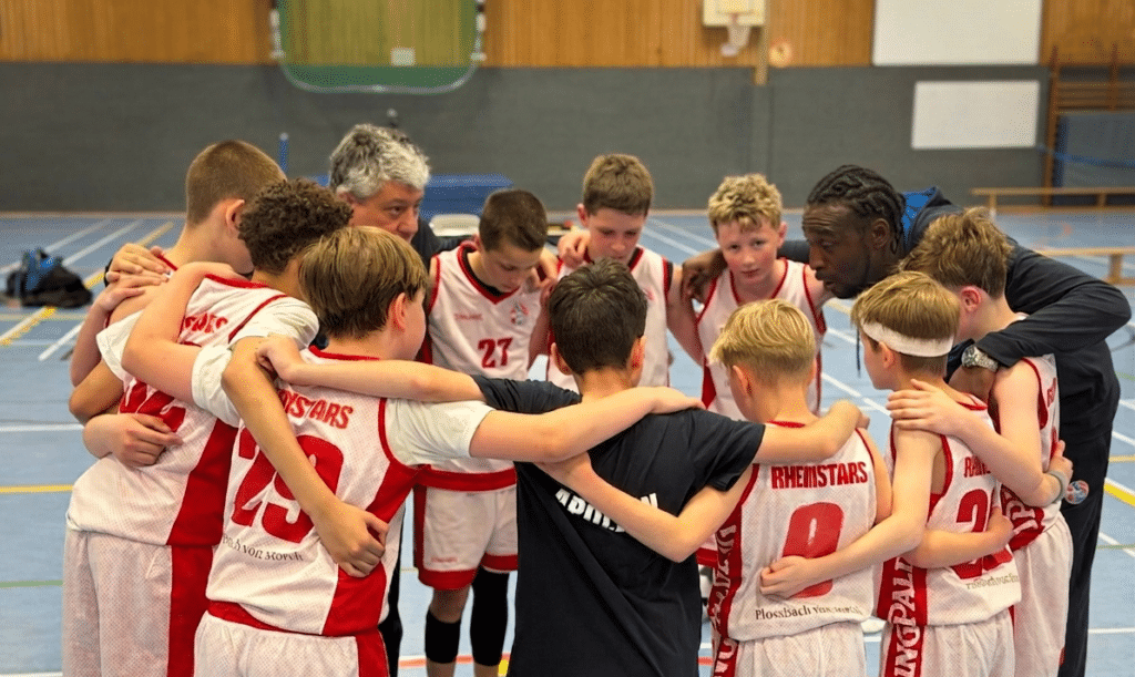 Basketballteam RheinStars Köln im Teamkreis mit Trainern in einer Sporthalle.
