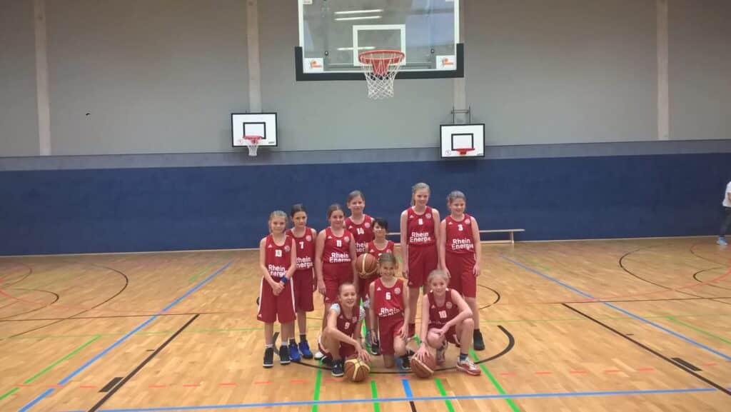 Mädchen-Basketballteam der RheinStars Köln in roter Trainingskleidung posiert in einer Sporthalle.