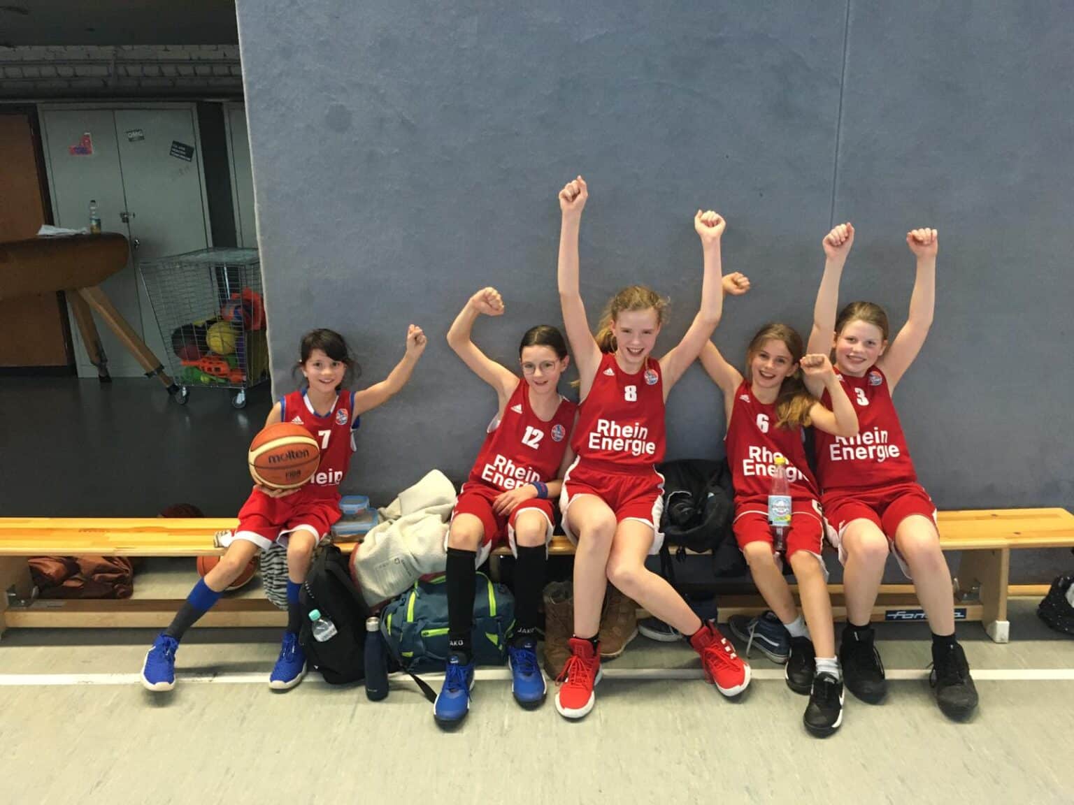 Fünf Basketballerinnen der RheinStars Köln in roten Trikots sitzen auf einer Bank und recken ihre Fäuste triumphierend in die Luft.