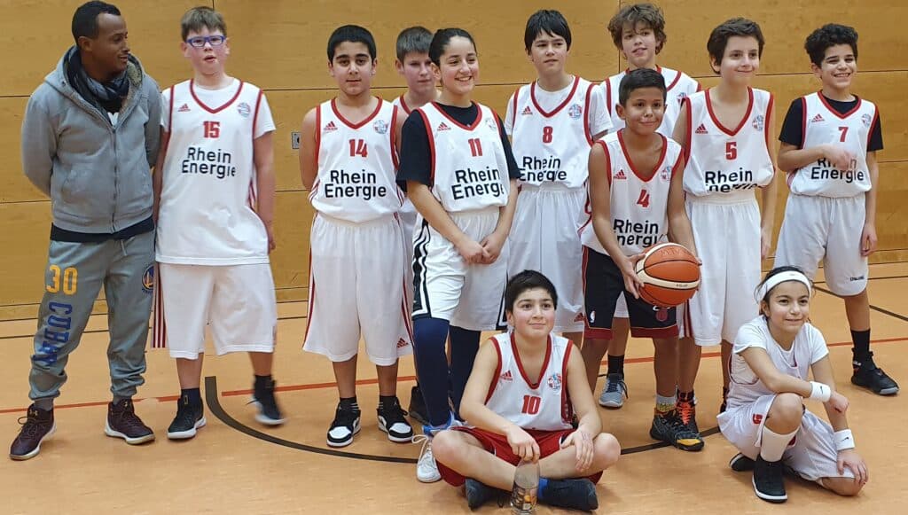 Jugendbasketballmannschaft RheinStars Köln posiert mit Trainer in der Sporthalle.