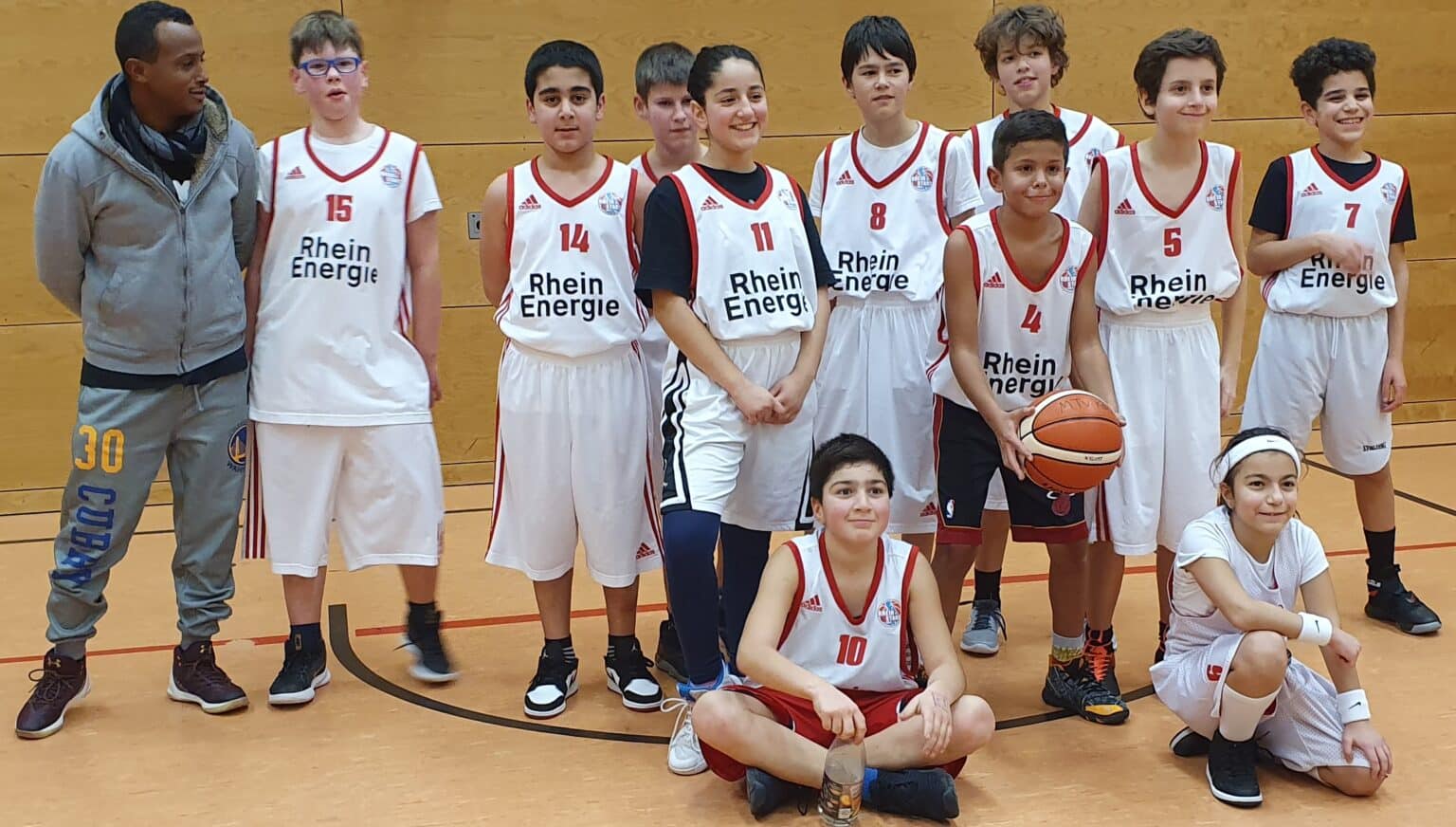 Jugendbasketballmannschaft RheinStars Köln posiert mit Trainer in der Sporthalle.