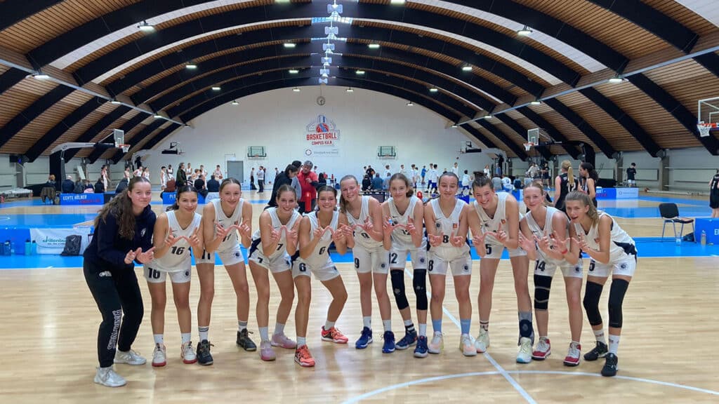 Frauenbasketballteam der RheinStars Köln posiert lächelnd in einer Sporthalle.