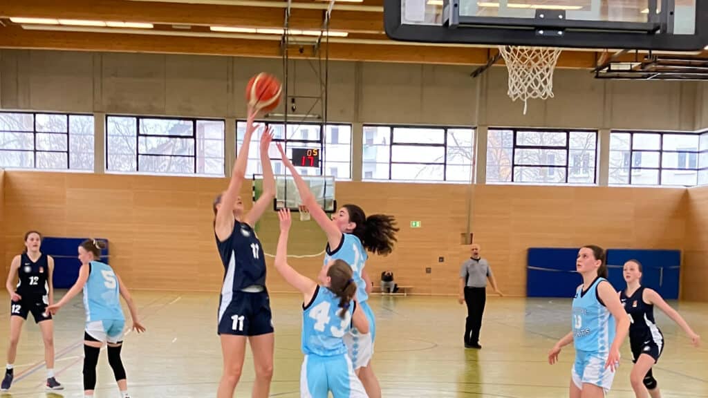 Basketballspiel der RheinStars Köln: Spielerin in dunklem Trikot wirft Ball auf Korb, Gegnerinnen in hellblauem Trikot verteidigen in der Sporthalle.