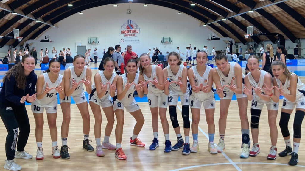 Frauenbasketballteam RheinStars Köln posiert lächelnd in Spielkleidung auf dem Basketballplatz.