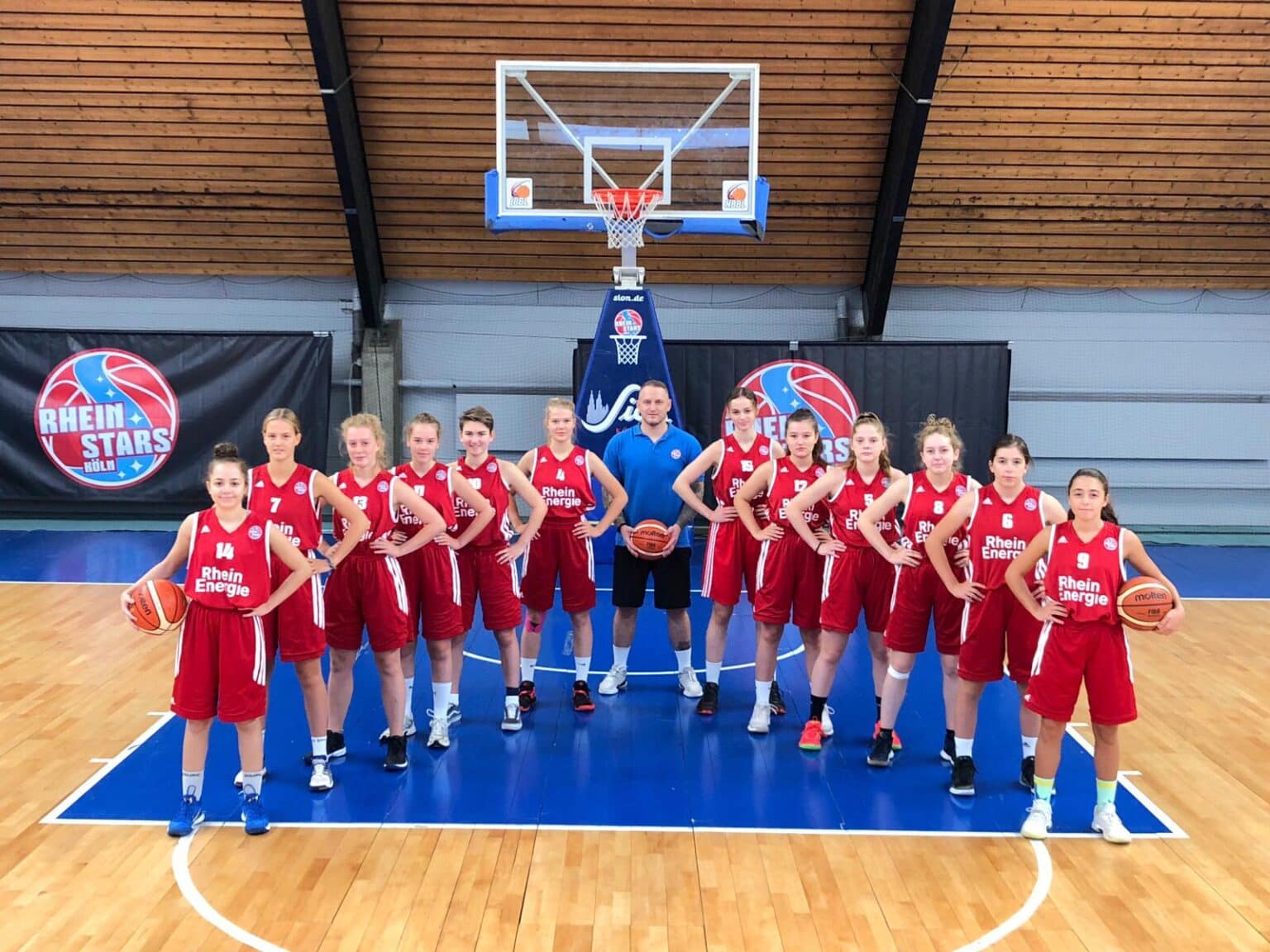 Mädchen-Basketballteam RheinStars Köln mit Trainer posiert in roter Sportkleidung in einer Sporthalle.