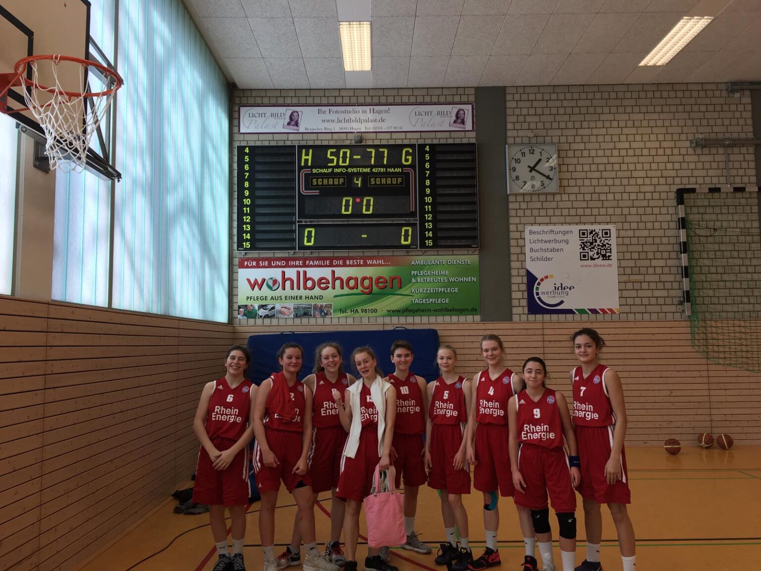 Zehn Basketballspielerinnen der RheinStars Köln in roten Trikots stehen in einer Sporthalle.