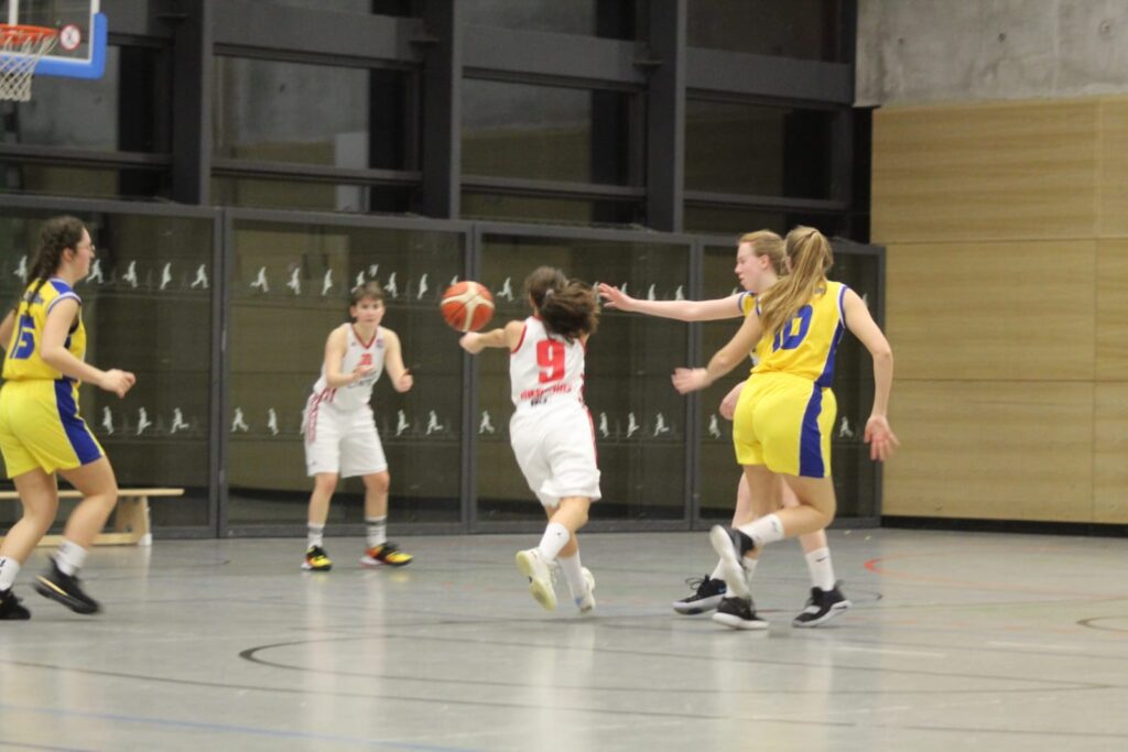 Basketballspiel der RheinStars Köln: Spielerin in Weiß mit Ball wird von Spielerinnen in Gelb-Blau verteidigt.