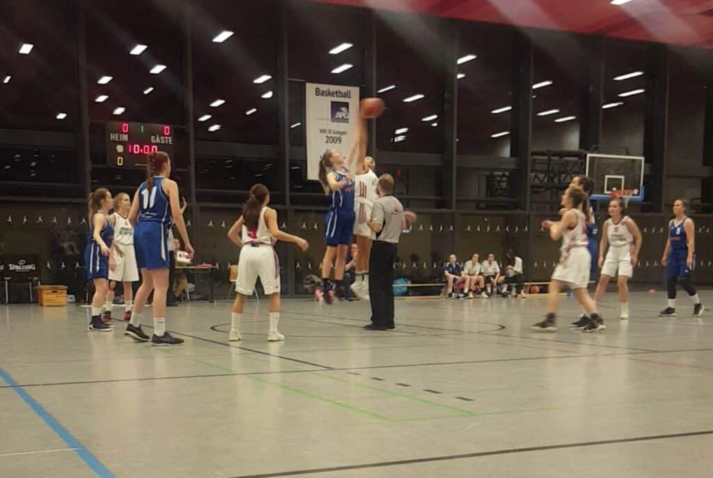 Basketballspiel der RheinStars Köln: Spielerin in blauem Trikot wirft Ball auf Korb, Gegnerinnen in weißen Trikots verteidigen.