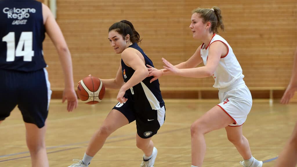Basketballspiel der RheinStars Köln: Spielerin in schwarzem Trikot dribbelt gegen Verteidigerin in weißem Trikot in einer Sporthalle.
