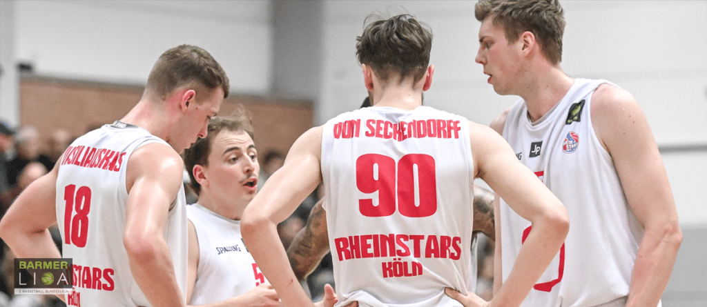 Basketballspieler der RheinStars Köln in weißen Trikots besprechen sich während eines Spiels in der Halle.
