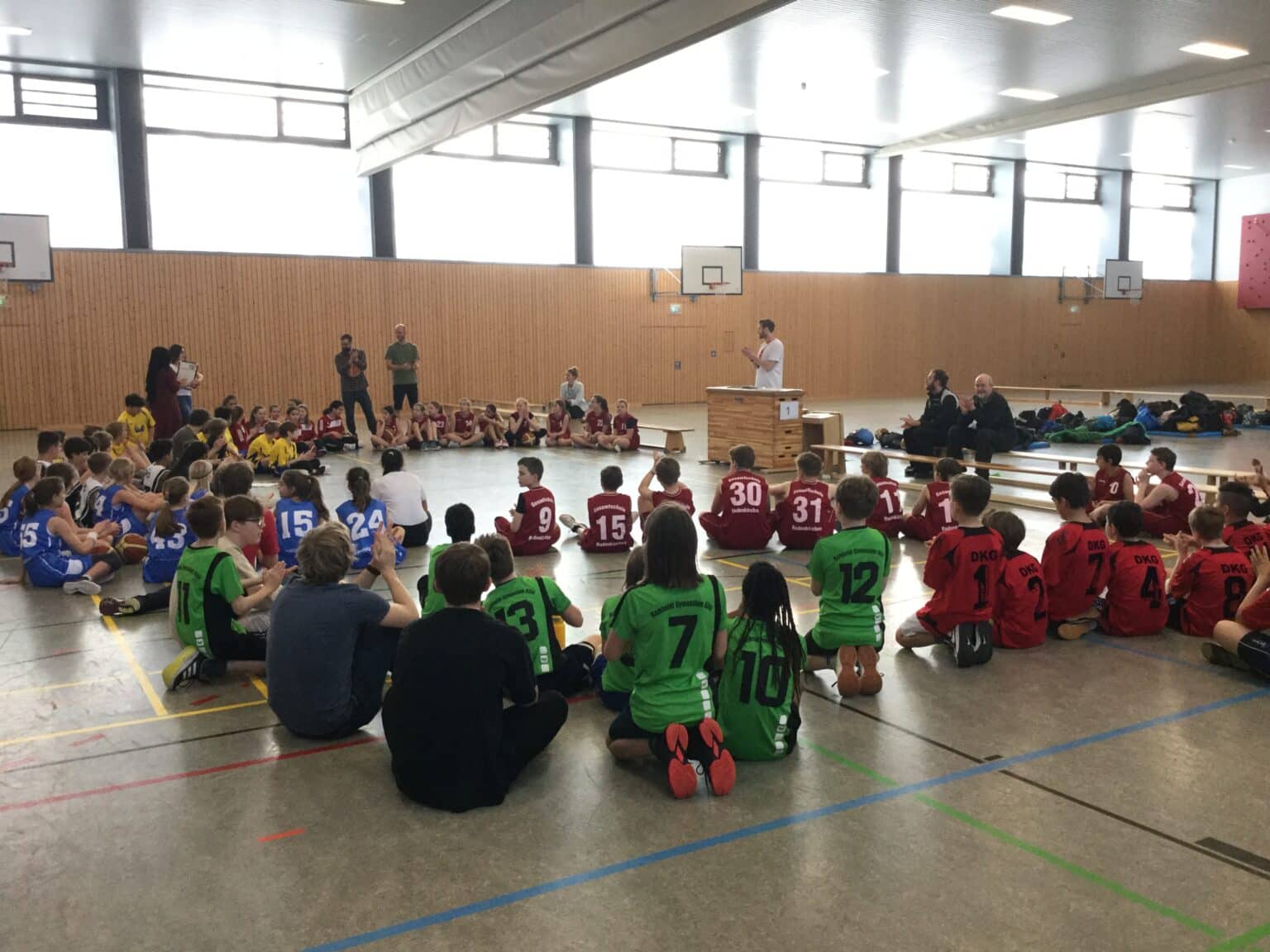 Kinder verschiedener Sportteams sitzen in einer Sporthalle und hören einem Trainer zu.