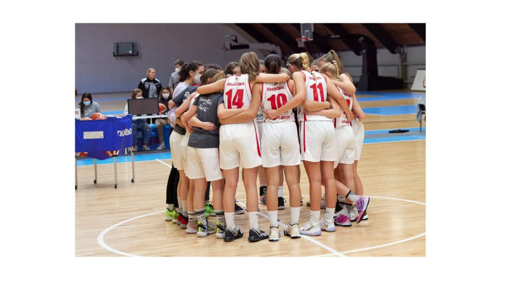 RheinStars Köln Basketballteam in Teamkreis auf Hallenboden vereint.