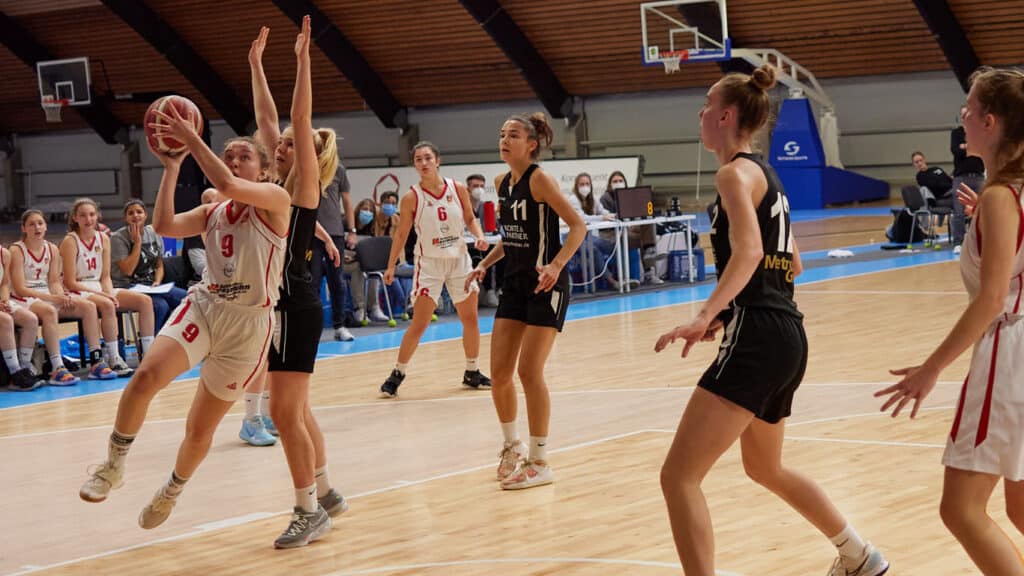 Basketballspiel der RheinStars Köln: Spielerin im roten Trikot wirft Ball, schwarze Gegenspielerinnen verteidigen in Sporthalle.