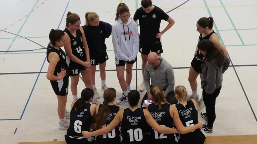 Trainingsszene der RheinStars Köln: Trainer bespricht Taktik mit weiblicher Basketballmannschaft in Sporthalle.