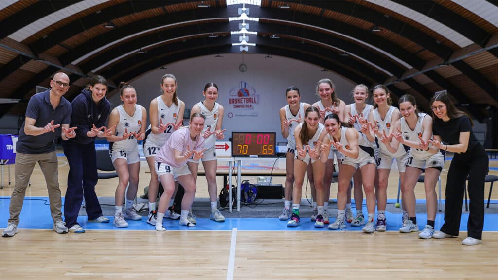 Basketballmannschaft RheinStars Köln posiert jubelnd in der Sporthalle vor dem Anzeigetableau.