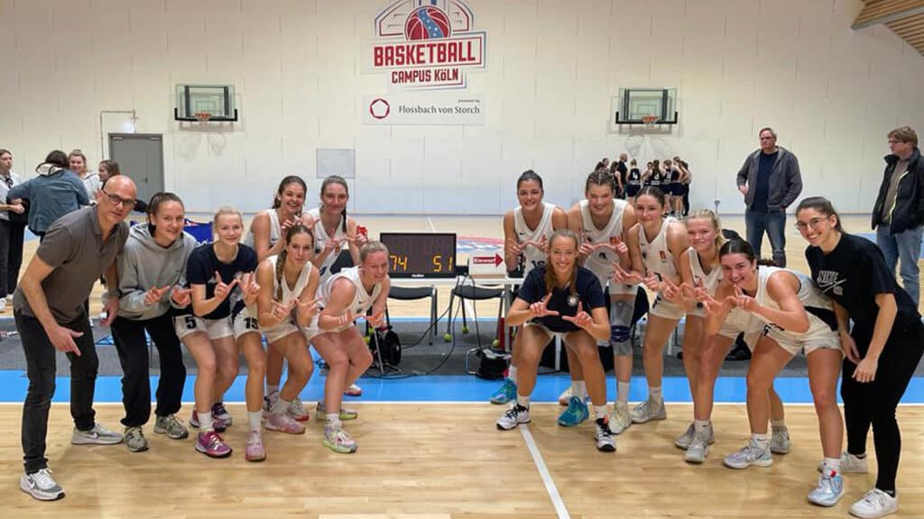 Frauenbasketballmannschaft der RheinStars Köln posiert lachend auf dem Spielfeld einer Sporthalle.