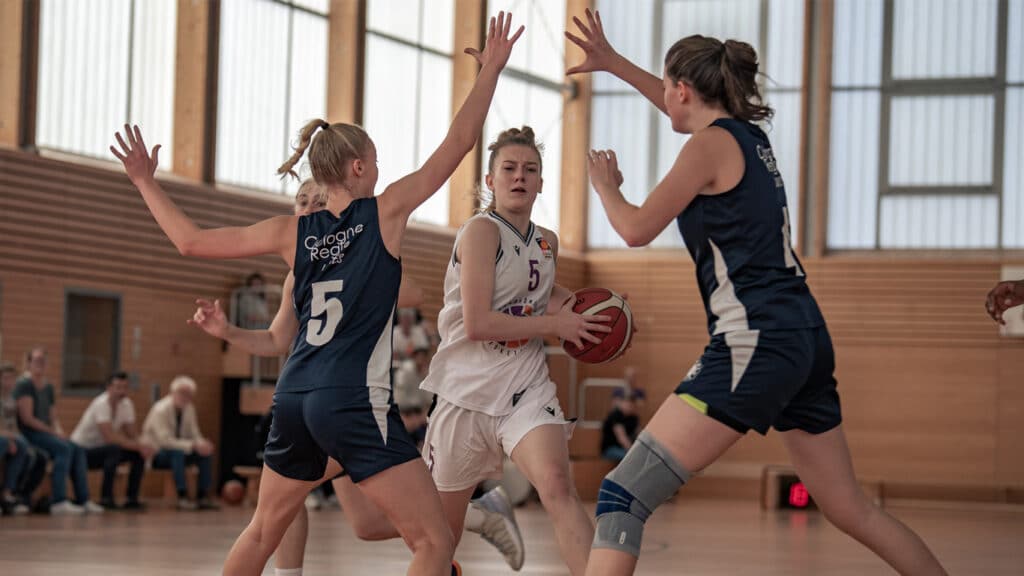 Basketballspiel der RheinStars Köln: Spielerin in weißem Trikot mit Ball umgeben von Verteidigern in dunklen Trikots.