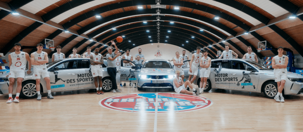 Basketball-Mannschaft RheinStars Köln posiert mit weißen Fahrzeugen in der Sporthalle.
