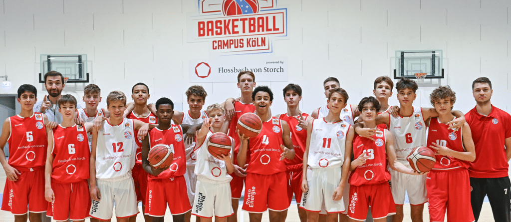 Basketballmannschaft RheinStars Köln posiert geschlossen mit Bällen vor weißer Hallenwand.