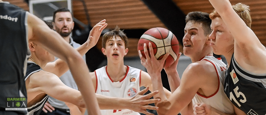 Basketballspiel der RheinStars Köln: Spieler in weißem Trikot mit Ball umgeben von Gegnern in schwarzen Trikots.
