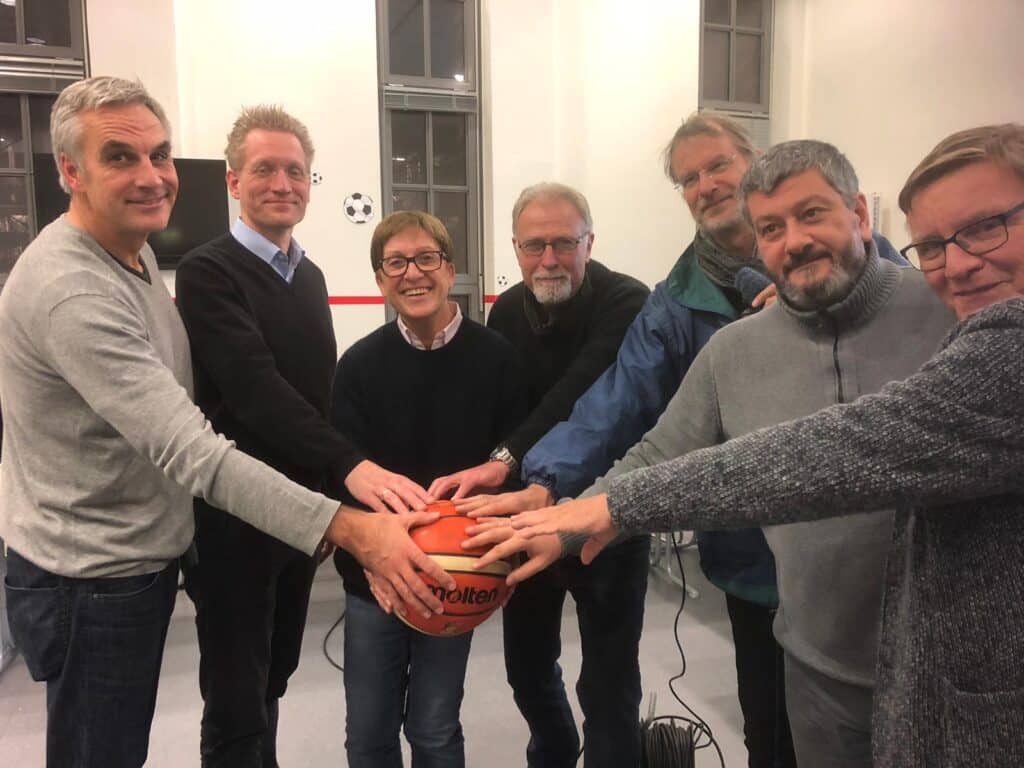 Sieben Personen der RheinStars Köln halten gemeinsam einen roten Volleyball in die Kamera.
