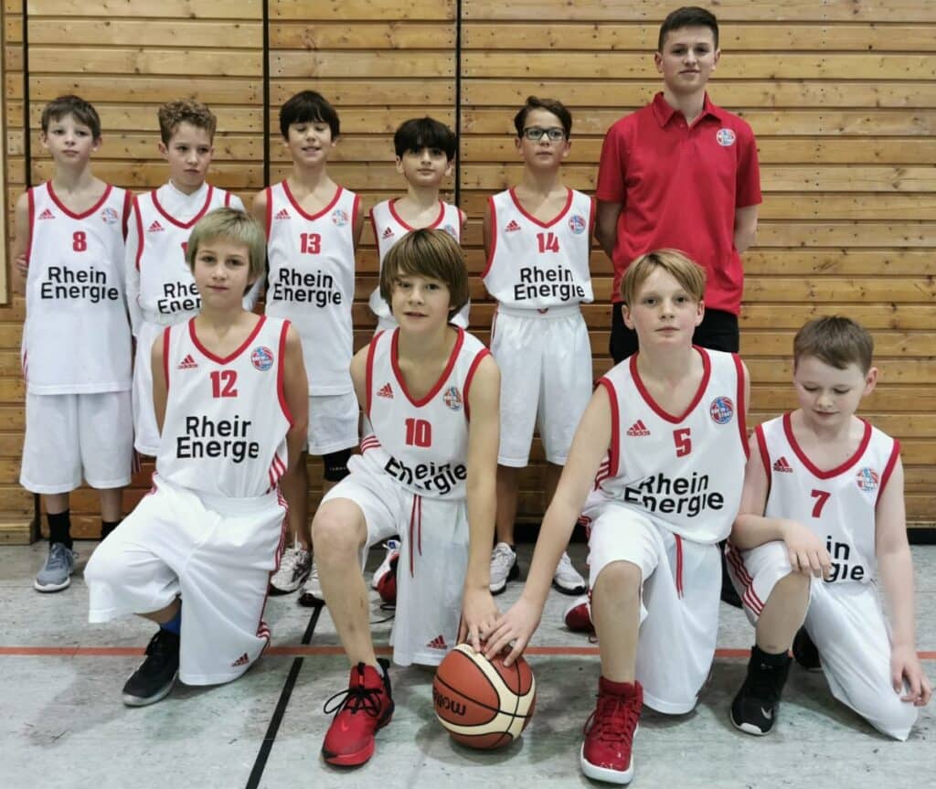 Jugendbasketballteam RheinStars Köln in weißen Adidas-Trikots mit Sponsor „Rhein Energie" posiert in einer Sporthalle vor Holzwand.