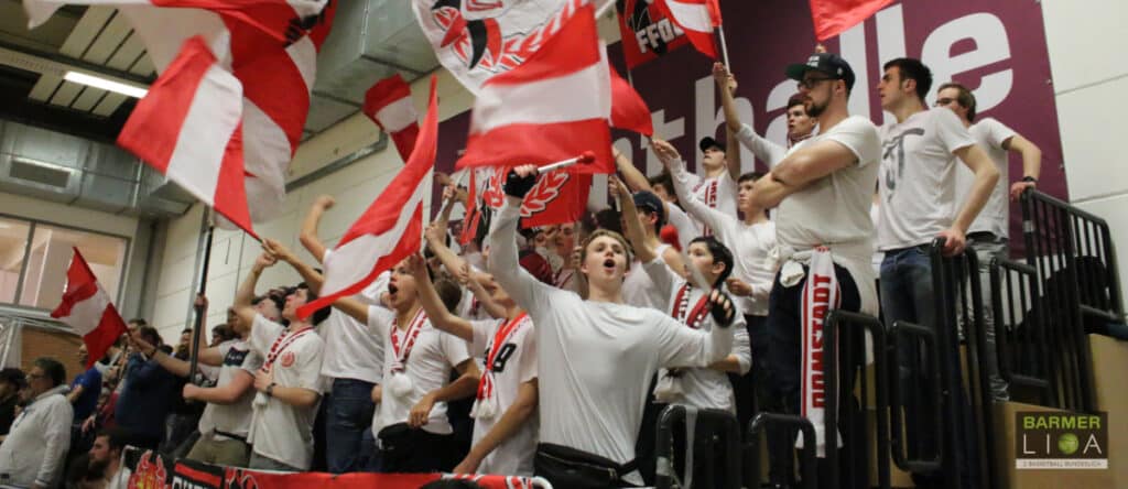 Fans der RheinStars Köln schwenken rot-weiße Flaggen in einer Sporthalle.