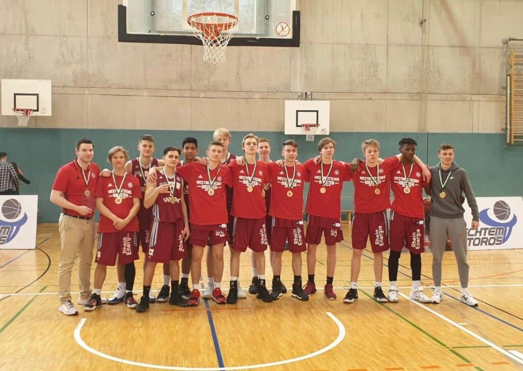 Jugendbasketballteam der RheinStars Köln mit Goldmedaillen in einer Sporthalle posierend.