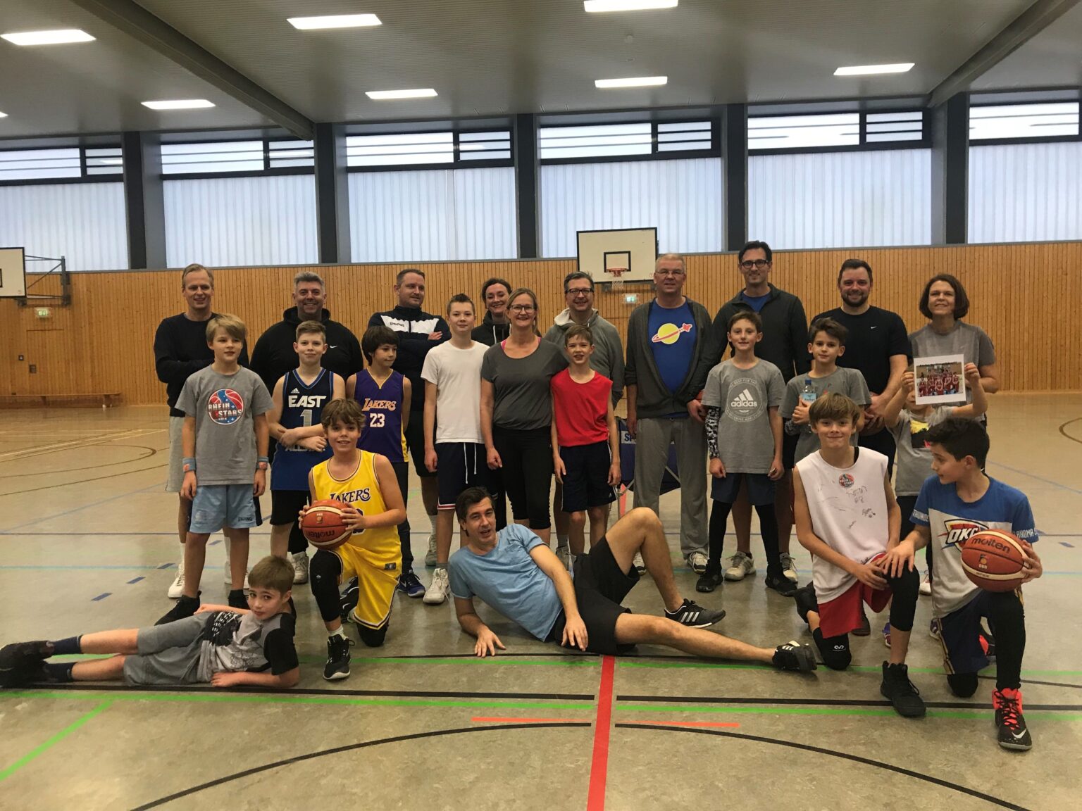 Gruppe von Kindern und Erwachsenen der RheinStars Köln posiert gemeinsam in einer Sporthalle mit Basketbällen.
