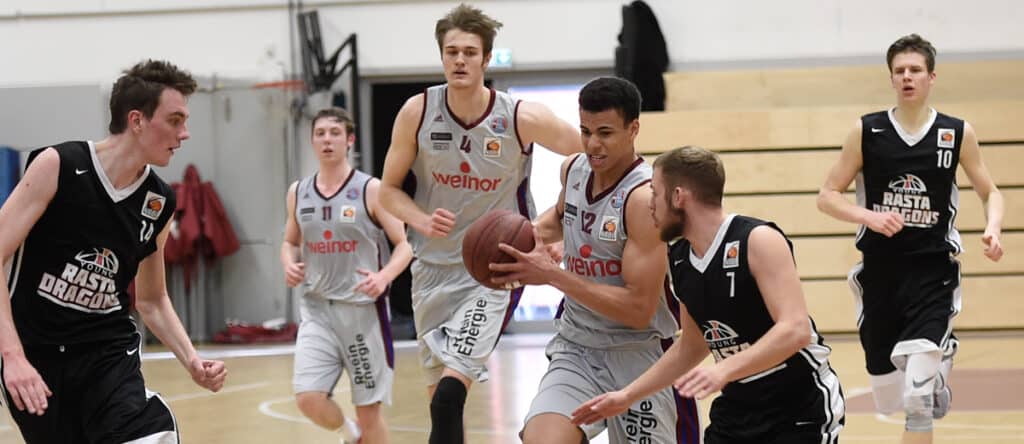 Basketballspiel der RheinStars Köln: Spieler in grauen Trikots mit "Welnor"-Sponsor gegen Gegner in schwarzen Trikots mit "RheinStars Dragons"-Logo in Sporthalle.