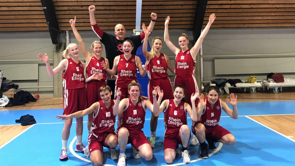 Basketballteam RheinStars Köln in burgunderroten Trikots feiert jubelnd in der Sporthalle.