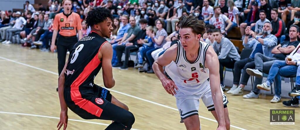Basketballspiel der RheinStars Köln: Spieler in weißem Trikot verteidigt gegen Gegner in schwarzem Trikot mit rotem Streifen.
