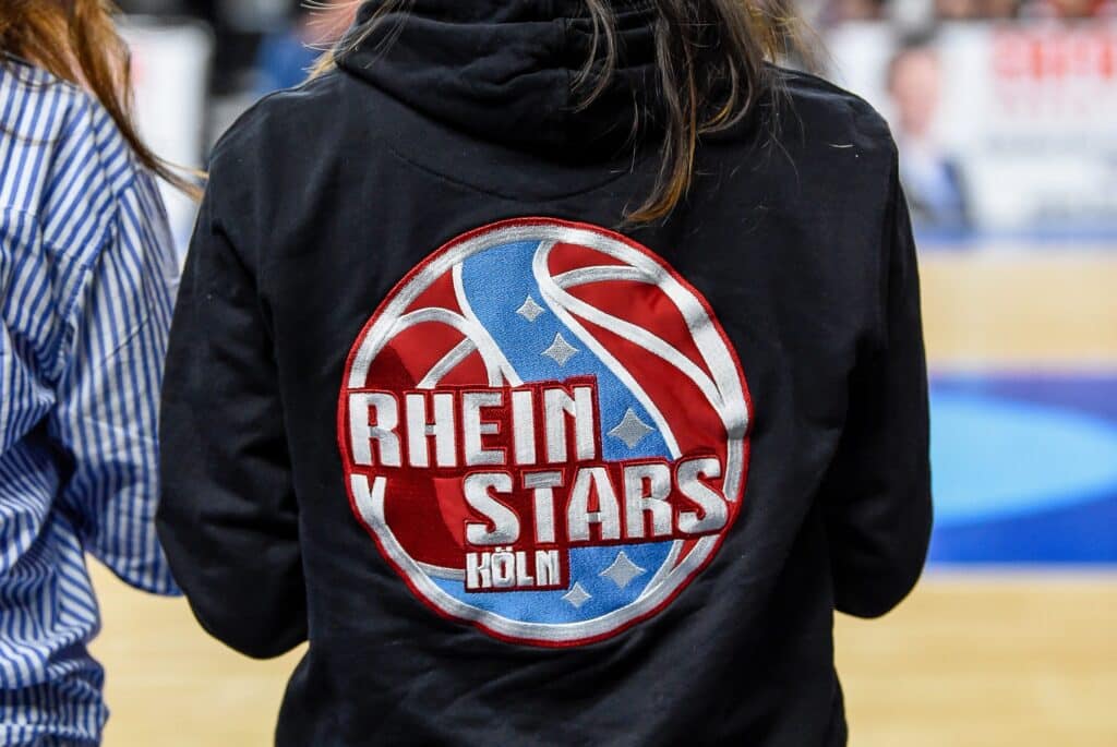 Person trägt schwarze Jacke mit RheinStars Köln Wappen und Logo.