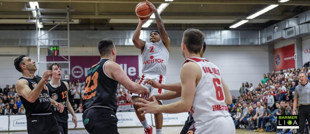 Basketballspiel der RheinStars Köln: Spieler in Weiß wirft von außen, schwarze Gegenspieler verteidigen.