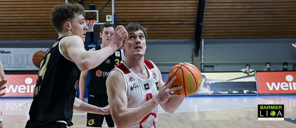 Basketballspiel: Spieler der RheinStars Köln in weißem Trikot mit Ball, verteidigt von Gegner in schwarzem Trikot.