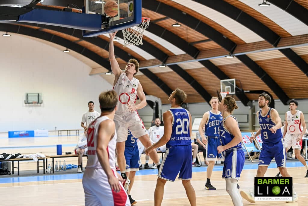 Basketballspieler der RheinStars Köln wirft Ball zum Korb, Gegner in bluen Trikots verteidigt.