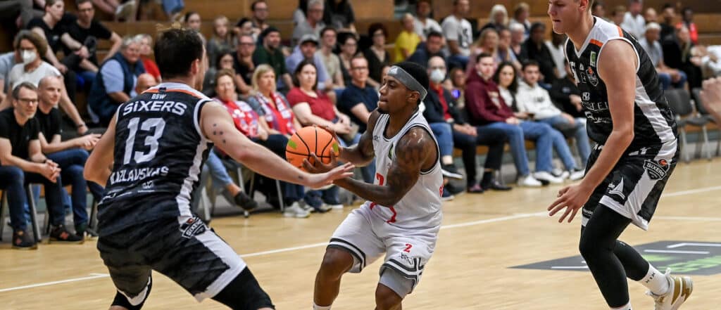 Basketballspiel der RheinStars Köln: Spieler in weißem Trikot dribbelt gegen Gegner in dunklem Sixers-Trikot.